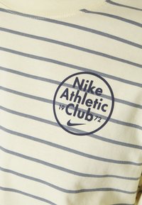 Randig mjuk bomullströja med korta ärmar i ljus krämfärg med marinblå ränder. Har en rund logotyp med "Nike Athletic Club 1972" i mitten.