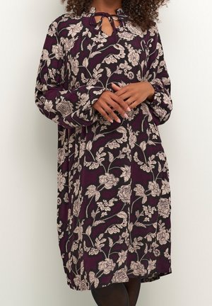 Woman wearing a dark purple dress with white floral patterns, long sleeves, and a tie closure at the neckline, standing with hands clasped.
