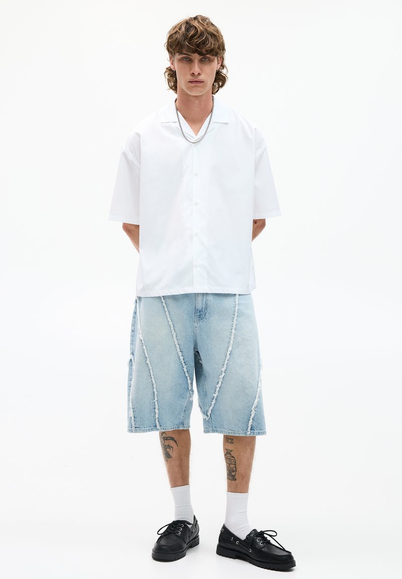 Camicia bianca a maniche corte con bottoni, pantaloni corti in denim chiaro con bordi sfrangiati e cuciture a contrasto, indossati con scarpe nere e calze bianche.