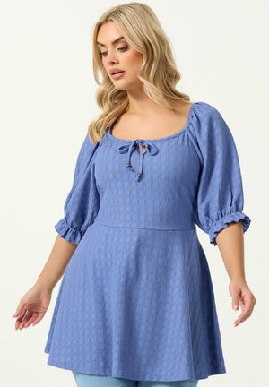 Kvinde med blondt hår iført en tekstureret blå kjole med oppustede ærmer og en bindedetalje ved den firkantede halsudskæring, der kigger nedad.