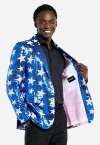 Paillette blazer in blauw met een wit sterrenpatroon, met een roze binnenvoering en een zichtbaar label aan de binnenkant. Model die glimlacht en een zwart shirt draagt.