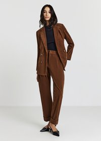 Brauner Cord-Anzug mit einem doppelt geknöpften Blazer mit goldenen Knöpfen und passenden, konisch zulaufenden Hosen, kombiniert mit einem schwarzen, gerippten Oberteil und schwarzen Schuhen.