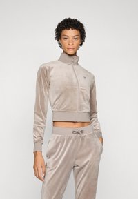 Cropped zip-up jacket made of soft beige velour, featuring a high collar, front pockets, and ribbed cuffs, paired with matching joggers.