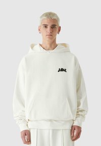 Biela mikina z bavlny s predným vreckom a čiernym logom na hrudi, s voľným strihom a kapucňou so šnúrkou.