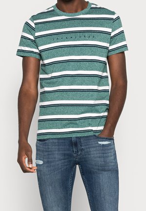 Camiseta de rayas verdes y blancas con mangas cortas, cuello redondo y un detalle de logo en el pecho. Llevada con jeans azules desgastados.
