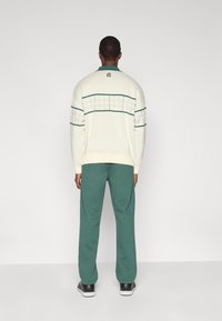 Λευκό πουλόβερ με πράσινες λεπτομέρειες, με σχέδιο πλέγματος στο στήθος, σε συνδυασμό με πράσινο-μπλε παντελόνι φόρμας και μαύρα παπούτσια.