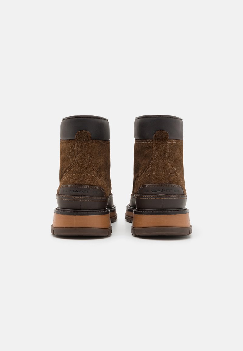 Bruine suède enkelboots met donkere leren accenten, een stevige rubberen zool en contrasterende stiksels. De achterkant toont het merklogo en textuurdetails.