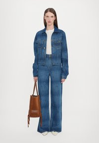 Denim jakna i široke hlače u tamnoplavoj boji s teksturiranim uzorkom. Model drži smešnu kožnu torbu s remenom za rame.