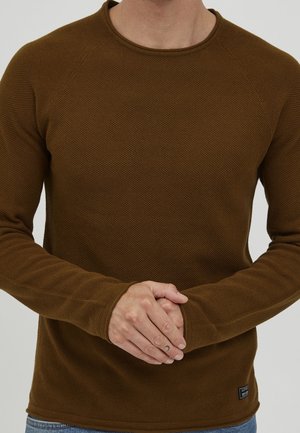 Mężczyzna w brązowej, teksturowanej, długorękawowej koszuli, z rękami splecionymi z przodu, widoczne jasnoniebieskie dżinsy na dole.