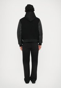 Hanorac negru cu mâneci din piele, având o croială lejeră și manșete ribbate. Asortat cu o pereche de pantaloni negri, văzut din spate.