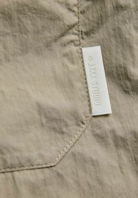 Material texturat bej, cu un buzunar cusut și o etichetă albă inscripționată cu "© JJXX STUDIO" atașată la cusătura buzunarului.