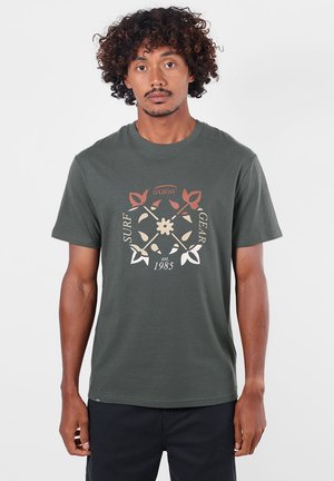 Homme aux cheveux bouclés portant un T-shirt vert foncé avec un motif floral et d’équipement de surf ainsi que le texte "Oxbow est. 1985" sur le devant, debout devant un fond blanc.
