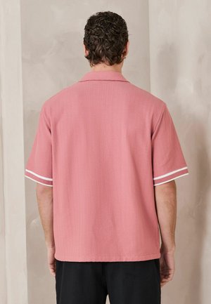 Man with curly dark hair wearing a pink short-sleeve shirt with white stripes on the cuffs, standing facing a beige wall.