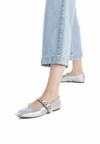 Bailarinas metálicas plateadas con un acabado texturado, que cuentan con una correa decorativa con tachuelas plateadas, combinadas con jeans cortos de color azul claro.