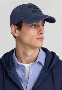 Cappellino in cotone blu scuro con testo ricamato "NEW YORK EAST COAST" sulla parte anteriore. Visiera curva, design casual, indossato con una felpa navy e una camicia a righe.