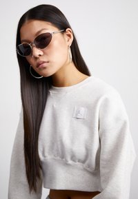 Grå, kort sweatshirt med et lite logo-patch. Oversized passform, myk tekstur. Slanke svarte solbriller med katteøyne-form og sølvfarget hardware.