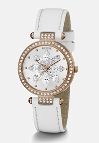 Ümmargune kell valge nahkrihmaga, roosakuldsetes aktsentides, kristallide kaunistustega ja valge näoga, millel on lillekujundusega ülemistel kristallidel. 