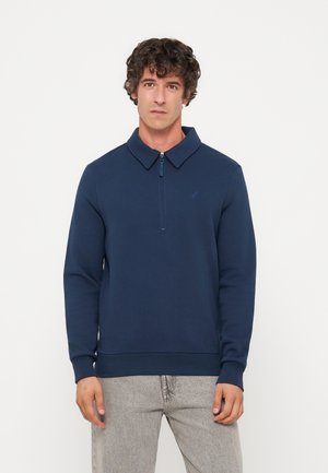 Homme aux cheveux bouclés portant un pull bleu marine à manches longues avec un col zippé et un pantalon gris clair, debout devant un fond uni.