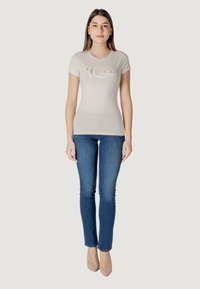 Camiseta ajustada color beige claro con un logo en cursiva de "Guess", combinada con jeans skiny azul oscuro y tacones nude. Fondo sencillo.