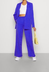 Costume violet ajusté avec un pantalon large et un blazer. Porté sur un crop top blanc. Assorti à des baskets blanches et un petit sac à main jaune.