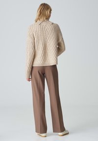 Beige Kabelstrickpullover mit Kragen, kombiniert mit hochgeschnittenen braunen Hosen und weißen Schuhen. Sanfte Textur und strukturierte Passform.