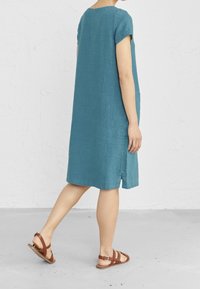Vestido de lino en color verde azulado con mangas cortas y aberturas laterales, de corte holgado, que llega hasta la mitad de la pantorrilla, combinado con sandalias marrones con tiras. Textura suave.