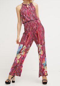 Vrouw in een levendige rode mouwloze jumpsuit met abstracte patronen en grote kleurrijke bloemenprints, gecombineerd met zwarte platformhakken.