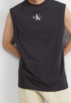 Mann trägt ein schwarzes ärmelloses Calvin Klein Jeans Shirt und beige Hosen, steht mit hinter dem Rücken verschränkten Händen vor einem weißen Hintergrund.