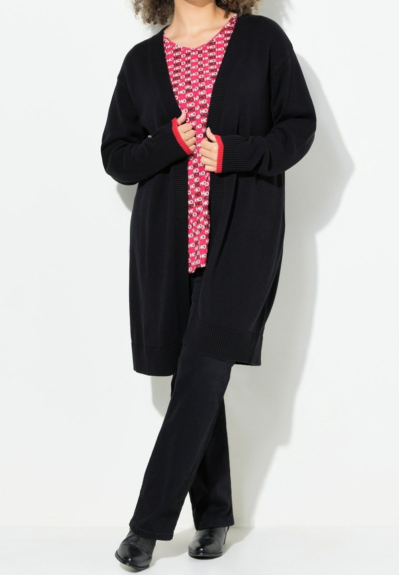 Femme portant un long cardigan noir par-dessus un haut à motifs rouges et blancs avec le texte "HO", un pantalon noir et des chaussures noires, debout contre un mur blanc.