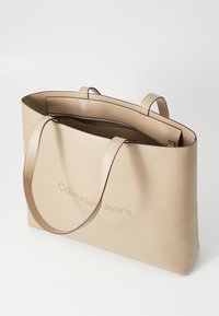 Bež kožna torba s dva ručka, zatvaranjem na patentni zatvarač i utisnutim logotipom "Calvin Klein Jeans" sprijeda. Glatka tekstura, minimalistički dizajn.