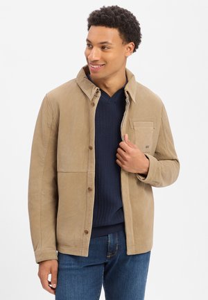 Man draagt een beige jas met knopen over een marineblauwe trui en een blauwe spijkerbroek, glimlacht en kijkt opzij tegen een witte achtergrond.