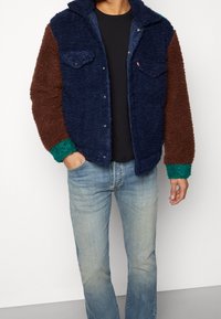 Chaqueta difusa con bloques de color en secciones azul, marrón y verde. Cuenta con un cuello, cierres a presión y dos bolsillos en el pecho. Textura suave.