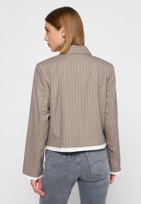 Bruine gestreepte cropped jas met een notch kraag, voorzien van een witte bies langs de zoom en de manchetten. De stof heeft een gladde textuur.