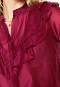 Burgundy blouse with ruffled neckline and button closure, made of sheer fabric, featuring a smooth texture and flowing design.