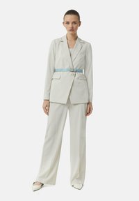 White blazer with structured shoulders and two front pockets, paired with wide-leg trousers and a light blue belt accentuating the waist.