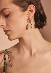 Guldhoopörhängen med en texturerad, pärlliknande hängande detalj, mot en mjuk, neutral bakgrund. Modellen bär en blommig linne med axelband.