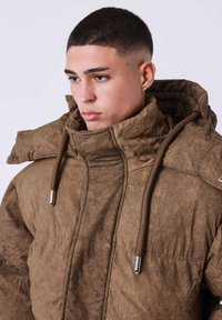 Chaqueta marrón texturizada con cuello alto, cordones ajustables y detalles metálicos. Presenta detalles de costura y capucha.