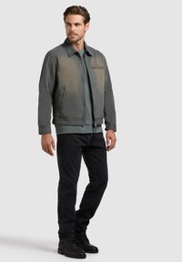 Egy férfi szürke cipzáros dzsekit, kékes-szürke inget, fekete farmert és fekete csizmát visel, kezei lazán mellett áll, és oldalra néz.