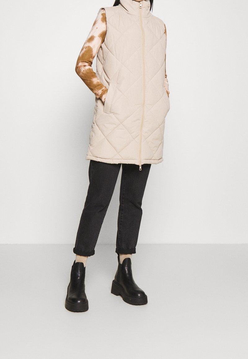 Gilet senza maniche trapuntato beige con colletto alto, zip frontale e tasche laterali, abbinato a pantaloni neri con risvolto e stivali neri robusti.