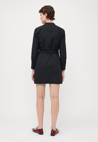 Abito nero a camicia con maniche lunghe, colletto e vita cintrata; texture liscia e design al ginocchio, abbinato a scarpe ballerine rosse.