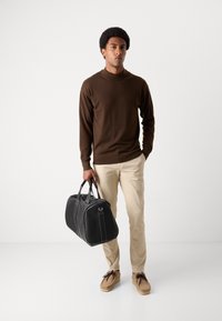 Zwarte leren reistas met twee bovenhandvaten en witte stiksels, gecombineerd met een bruine coltrui en beige chino's, gedragen met bruine schoenen.