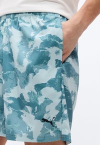 Χέρι στην τσέπη, φορώντας σορτς Puma με σχέδιο καμουφλάζ σε πετρόλ και λευκό, με ελαστική μέση και μαύρο λογότυπο Puma κοντά στο τελείωμα.