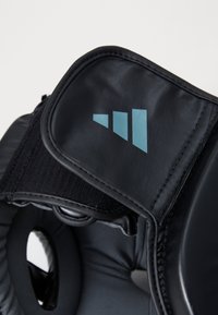 Gant de boxe noir rembourré avec un extérieur synthétique lisse, présentant un logo triangulaire bleu clair sur le poignet et une sangle de poignet ajustable.