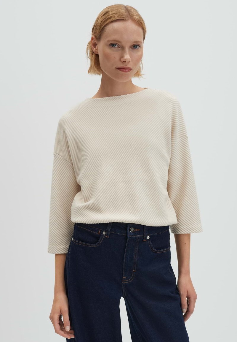 Donna che indossa un maglione beige a coste con maniche a tre quarti e jeans a vita alta blu scuro, in piedi davanti a uno sfondo semplice.