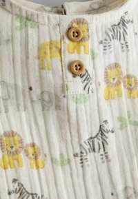 Gros plan sur un tissu blanc avec des imprimés légers d'animaux comprenant des lions, des zèbres et des girafes, agrémenté de deux petits boutons en bois sur une patte de boutonnage.