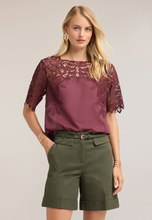 Lyshåret kvinde iført en lilla top med blondedetaljer og olivengrønne bælteshorts poserer med hænderne i lommerne mod en neutral baggrund.