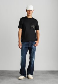 T-shirt preta com um bolso no peito, jeans azuis justos, e sapatilhas brancas, acompanhados de um boné branco. Tecido suave e ajuste casual.