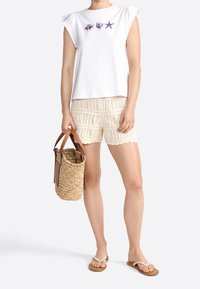Top blanco de manga corta con bordado de vida marina en morado, shorts de ganchillo beige y un bolso de mimbre. Sandalias de color claro complementan el conjunto.