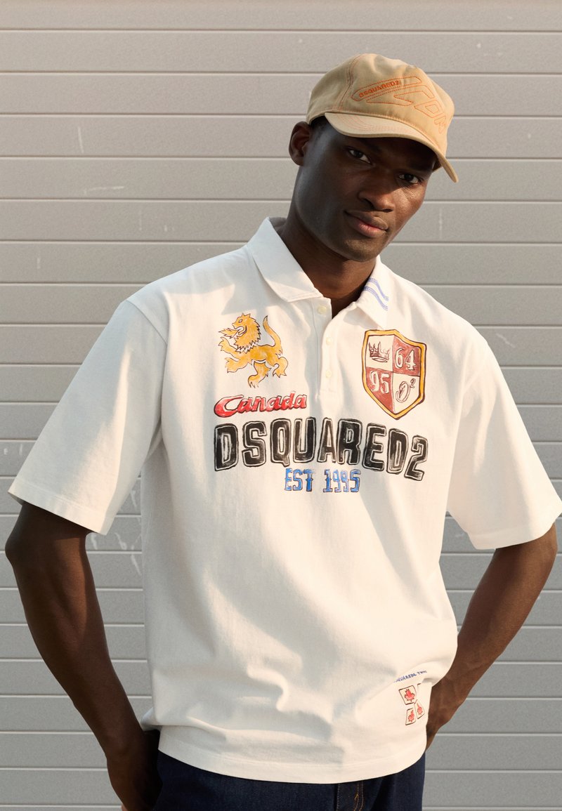 Tricou polo alb cu broderie cu leu, logo și anul 1995. Dispune de un guler cu trei nasturi, mâneci scurte și accente albastre dungi pe guler.