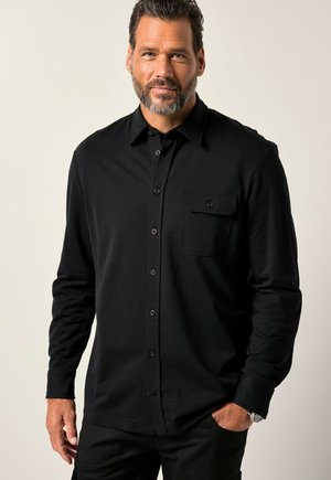 Camisa negra de manga larga con cuello abotonado, bolsillo frontal y textura sutil. Los botones contrastan en color. Se lleva con pantalones oscuros.
